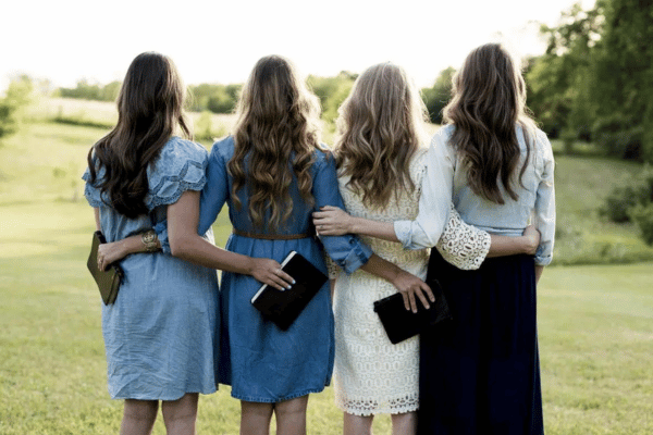 quatro mulheres de cabelos longos viradas de costas, num ambiente gramado a céu aberto, abraçadas, segurando bíblias, com vestidos na altura do joelho